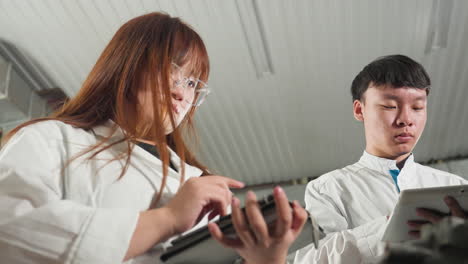 technicians in lab coats take notes on tablets while observing mechanical work in an automotive workshop, industrial setting with tools, equipment, and vehicles in the background