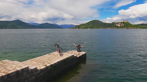 back view of happy woman with arms open and bicycle on concrete jetty edge of maggiore lake, italy