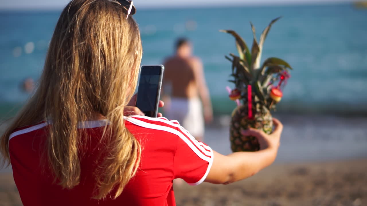 una turista con cabello rubio disfruta tomando una foto de un refrescante cóctel de piña en la playa en turquía - plano medio