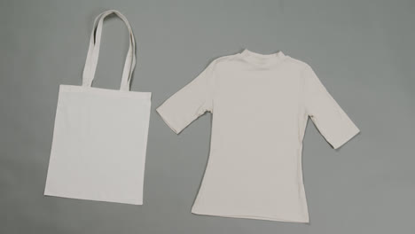 Close-up-of-white-bag-and-t-shirt-on-grey-background,-with-copy-space,-slow-motion