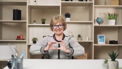 old-charismatic-woman,-looking-at-the-frame,-waves-her-hand-and-shows-a-heart