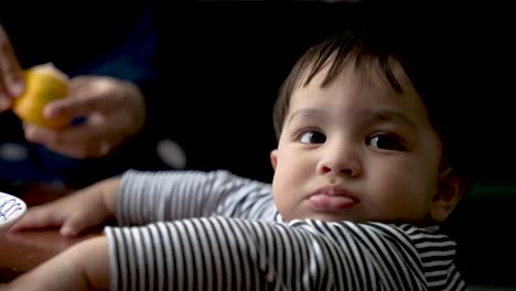 Süßes-Baby,-Das-Am-Tisch-Steht,-Mit-Einer-Person,-Die-Orange-Im-Hintergrund-Schält