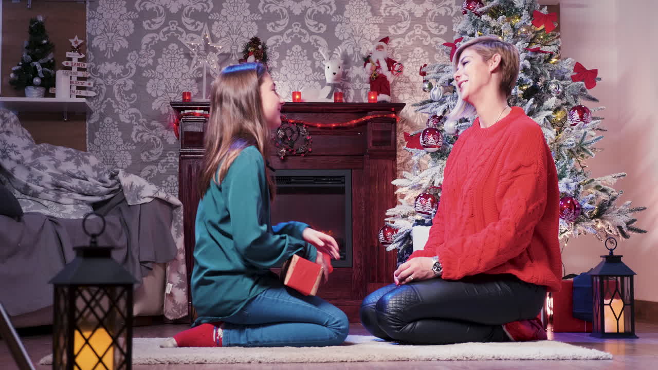 A mother and daughter exchange Christmas gifts by the fireplace