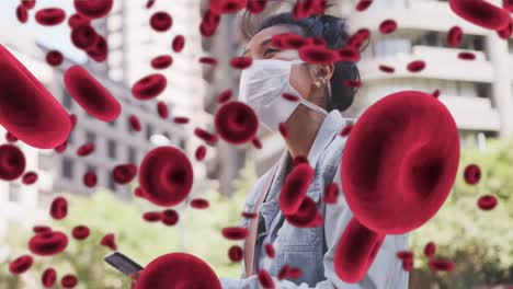 animation of floating blood cells over woman in face mask using smartphone in city