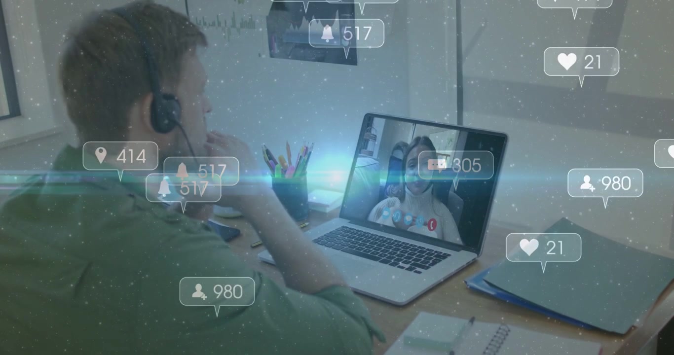Uomo che ascolta, con maglietta verde e cuffie, alla scrivania mentre una donna parla, con il laptop che mostra notifiche