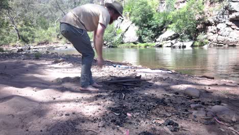 a bushman in an akubra hat stacks fire wood on a river bank in australia high country