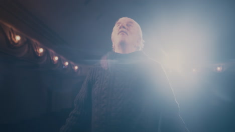 senior actor performing on stage tracking shot of aged man in sweater speaking and gesticulating while turning around in spotlight on stage during performance in dark theater