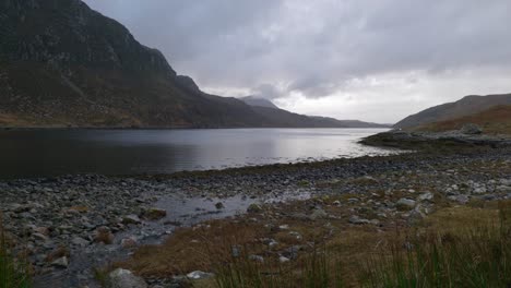 Bei-Ebbe-Im-Schottischen-Hochland-Fließt-Wasser-Sanft-In-Einen-See,-Während-Stimmungsvolle-Dunkle-Wolken-Hinter-Einem-Berg-über-Den-Himmel-Ziehen