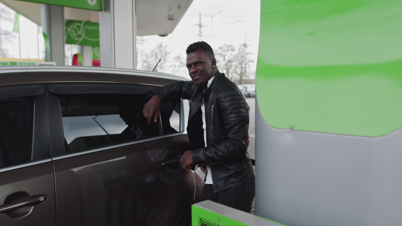 Portrait of an african guy at a gas station with coffee in hands refueling the car while traveling