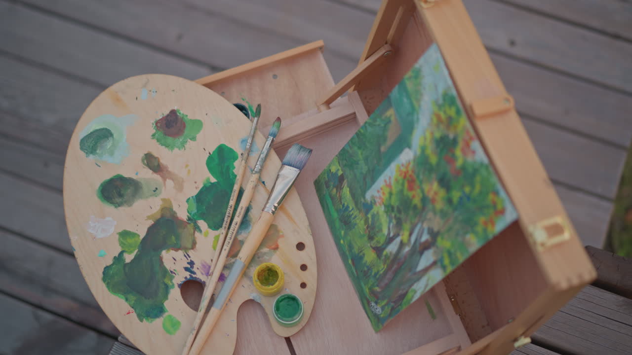 Palette in scatola di legno e piccola area di lavoro che mostra un paesaggio dipinto con un albero verde, pennelli appoggiati sulla palette, pozzetti di miscelazione pieni di pigmento verde, custodia portatile per cavalletto su ponte di legno, illuminato dal sole