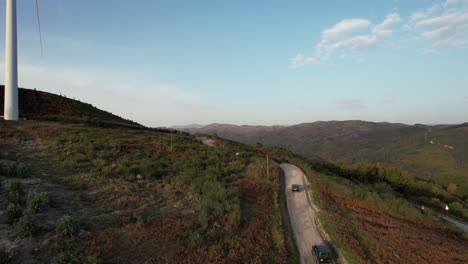 Seguimiento-De-Drones-A-Cuatro-Viajeros-En-Jeep-Conduciendo-Por-Una-Carretera-De-Molinos-De-Viento-Con-Paisaje-Montañoso