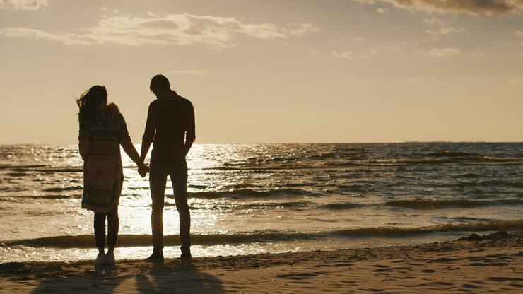 Silhouettes d'un jeune couple admirant le coucher de soleil sur la mer, s'enlaçant. Journée fraîche, le vent souffle