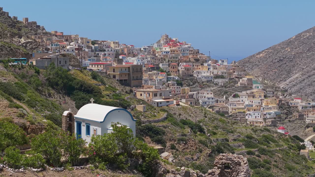 Drone shot reveals a whitewashed chapel in the foreground with the colorful traditional village of Olympos, Karpathos, in the background, set in a hillside landscape