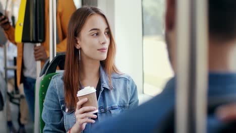 Jovencita-Hermosa-Sentada-En-El-Tranvía-Mientras-Va-A-Alguna-Parte,-Sosteniendo-Una-Taza-Para-Ir-Y-Bebiendo-Café-Caliente-Mientras-Sonríe-Y-Coquetea-Con-El-Chico-Que-Tiene-Delante