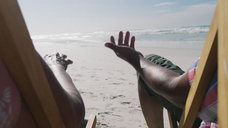 senior african american couple sitting on sunbeds and holding hands at the beach