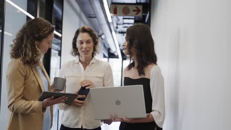 diversas compañeras en discusión usando computadora portátil y tableta en el pasillo de la oficina, cámara lenta