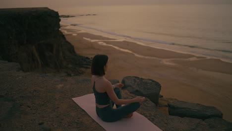 woman meditating on a cliff above the beach at sunset