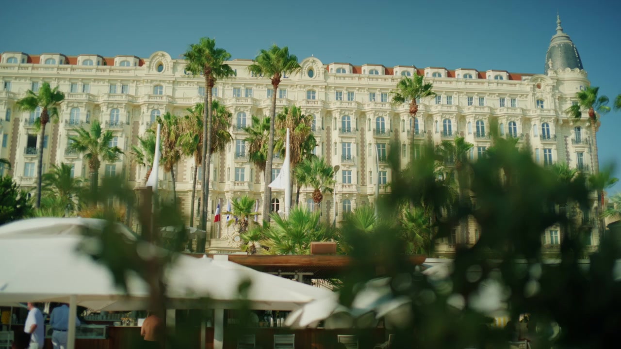 Ampia vista della facciata del Carlton Hotel incorniciata da palme e ombrelloni, Cannes, Francia