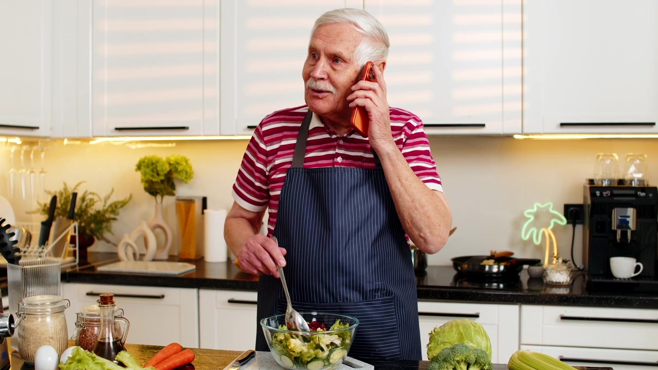 Älterer Mann mischt frischen Salat in der heimischen Küche, während er am Smartphone über einen gesunden Ernährungsplan spricht.