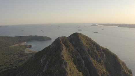 Slow-pivot-around-a-summit-of-a-pyramid-shaped-mountain-with-reveal-of-a-heart-shaped-bay-in-the-background-with-ships-at-the-anchorage,-on-the-sunset