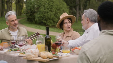 Middle-Aged-Man-Plating-A-Guitar-Sitting-At-Table-And-Talking-With-His-Friends-During-An-Outdoor-Party-In-The-Park