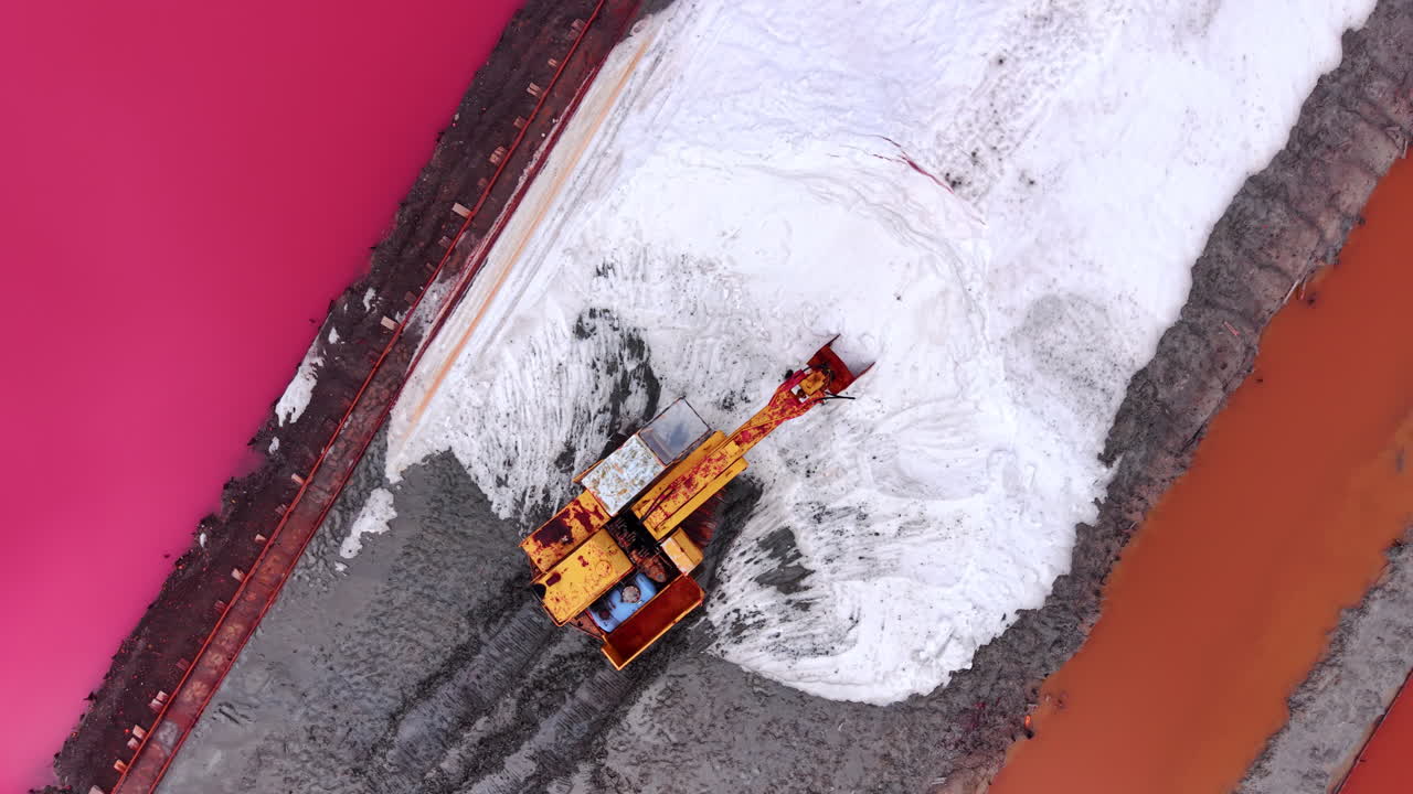 Escavatore giallo che scava in un grande cumulo di sale bianco accanto all'acqua rosa. Veduta aerea direttamente dall'alto di un escavatore che raccoglie sale.