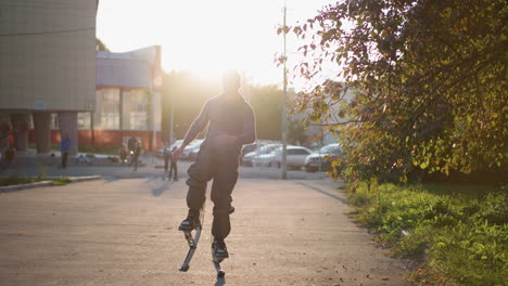 front shot of man wearing gray hoodie and dark gray pants jumping on spring stilts on paved road in residential area surrounded by houses during sunset, with serious expression