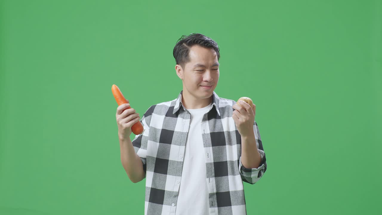 아시아 남성 농부 는 스튜디오 의 초록색 스크린 배경 에 따뜻 한 미소 를 지으면서 감자 와 당근 을 들고 있다