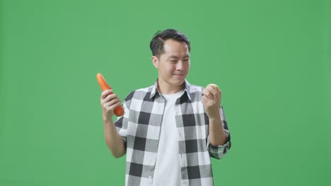 아시아 남성 농부 는 스튜디오 의 초록색 스크린 배경 에 따뜻 한 미소 를 지으면서 감자 와 당근 을 들고 있다