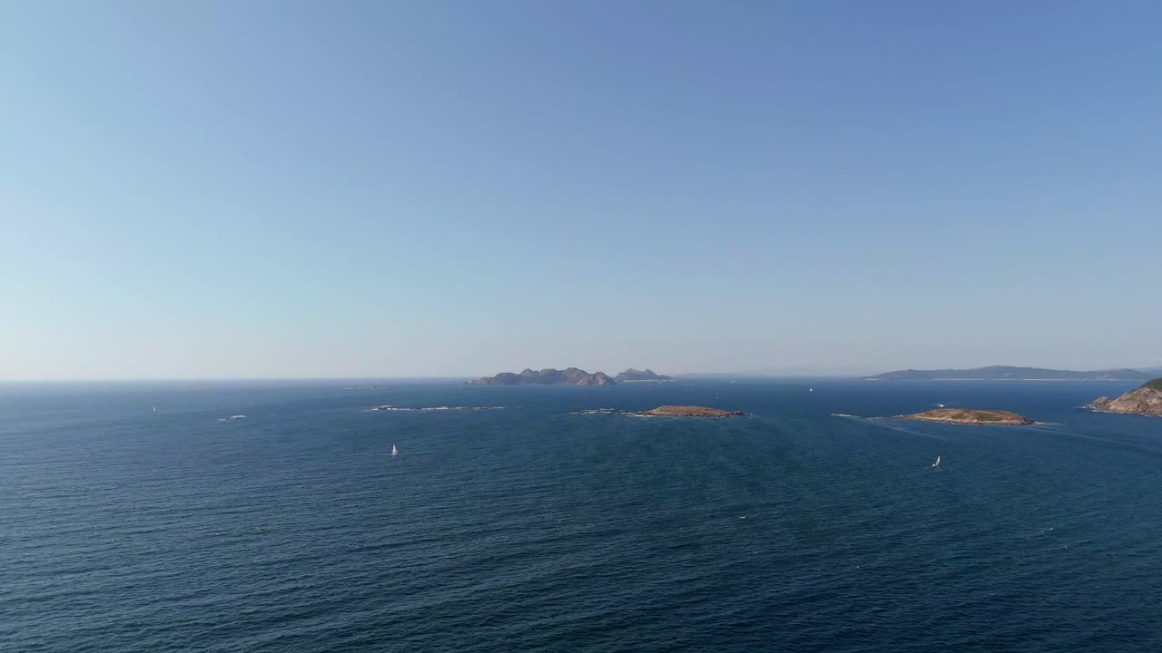 Aerial - 2 sailboats sailing past the horizon with Cíes Islands visible in the background.