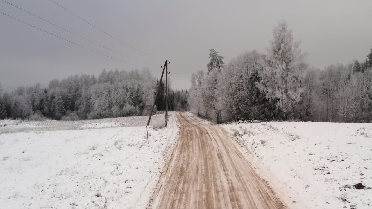 Aerial flight above gravel road on a cloudy winter day along powerlines. Snow covered ground and trees. Frozen forest after extreme cold.