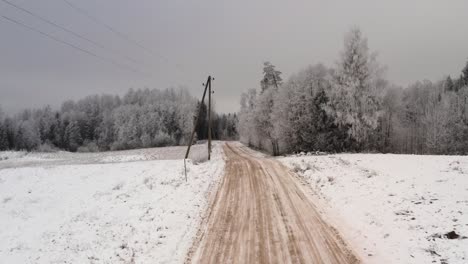 aerial flight above gravel road on a cloudy winter day along powerlines. snow covered ground and trees. frozen forest after extreme cold.