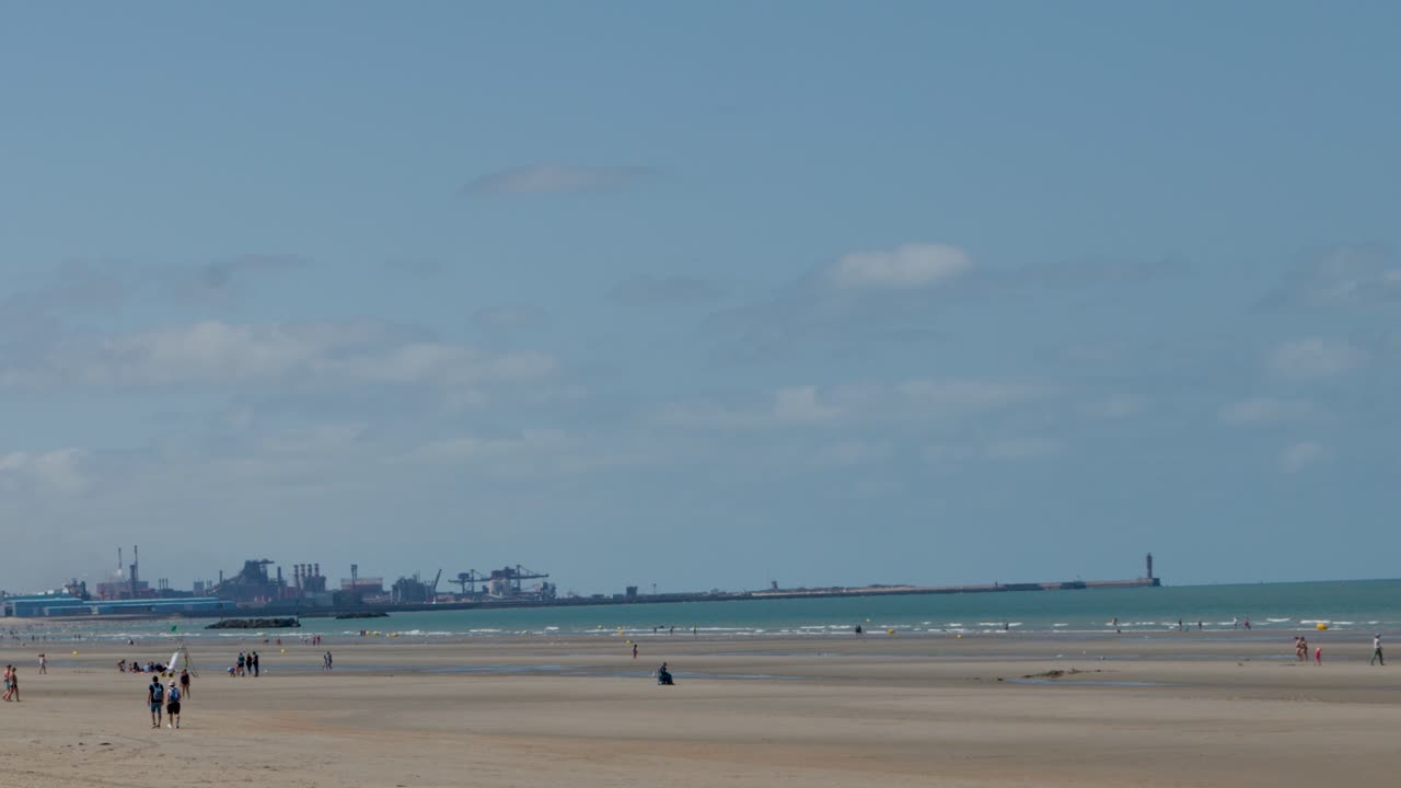 Groups stroll sandy shore with industrial harbor in background, under sunny skies, wide steady shot