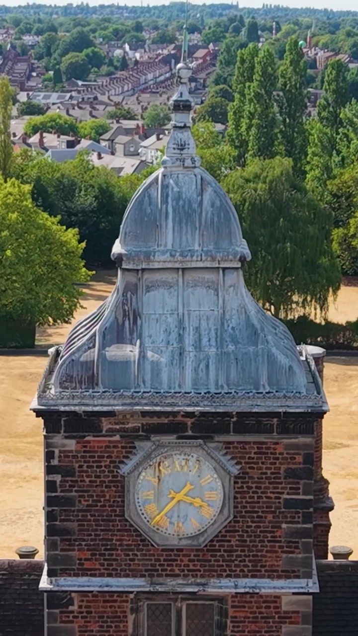 Aston Hall Jacobean clock tower in a locked descending drone shot, creating parallax as dry, drought-scorched lawns and tree-lined paths slide behind the historic roofline. Vertical format.