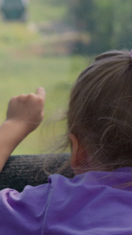 niña rubia con trenzas apunta a la aproximación del teleférico sentado en la ventana de la cabaña de la cuerda encima de la claridad del bosque en un día soleado cerca de la vista trasera en cámara lenta