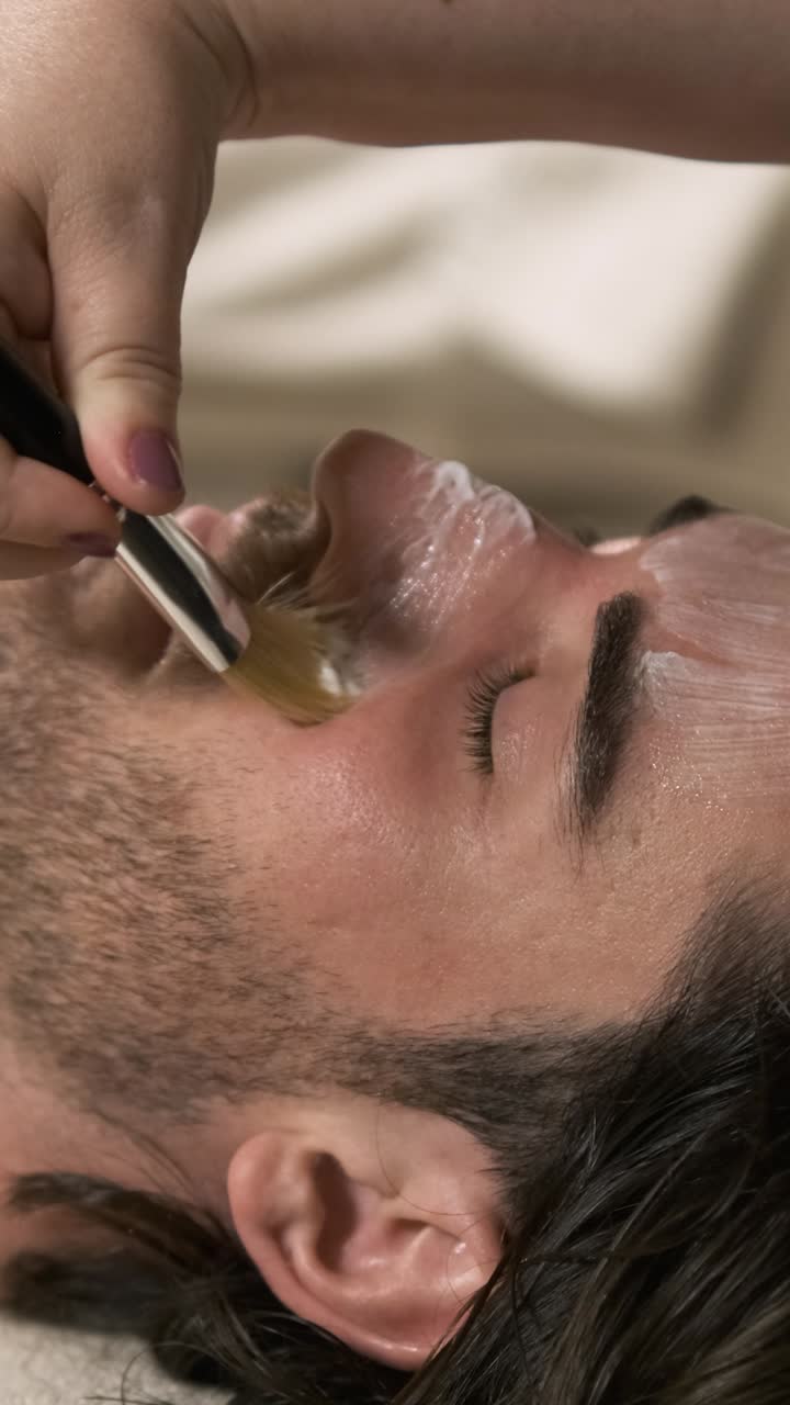Skilled beautician applying cream mask on relaxed man's face. Vertical