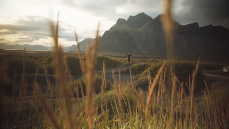 Wanderer-stands-in-the-distance-on-a-black-sanded-dune-and-spreads-his-arms-in-a-grassy-mountain-landscape