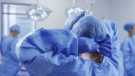 Mixed-race-female-surgeon-putting-on-protective-clothes-standing-in-operating-theatre