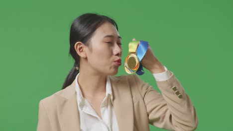슈트를 입은 아시아 사업가 여성이 손에 금메달을  자부심을 느끼며 스튜디오의 녹색 스크린 배경에서 첫 번째 우승자로 승리합니다.