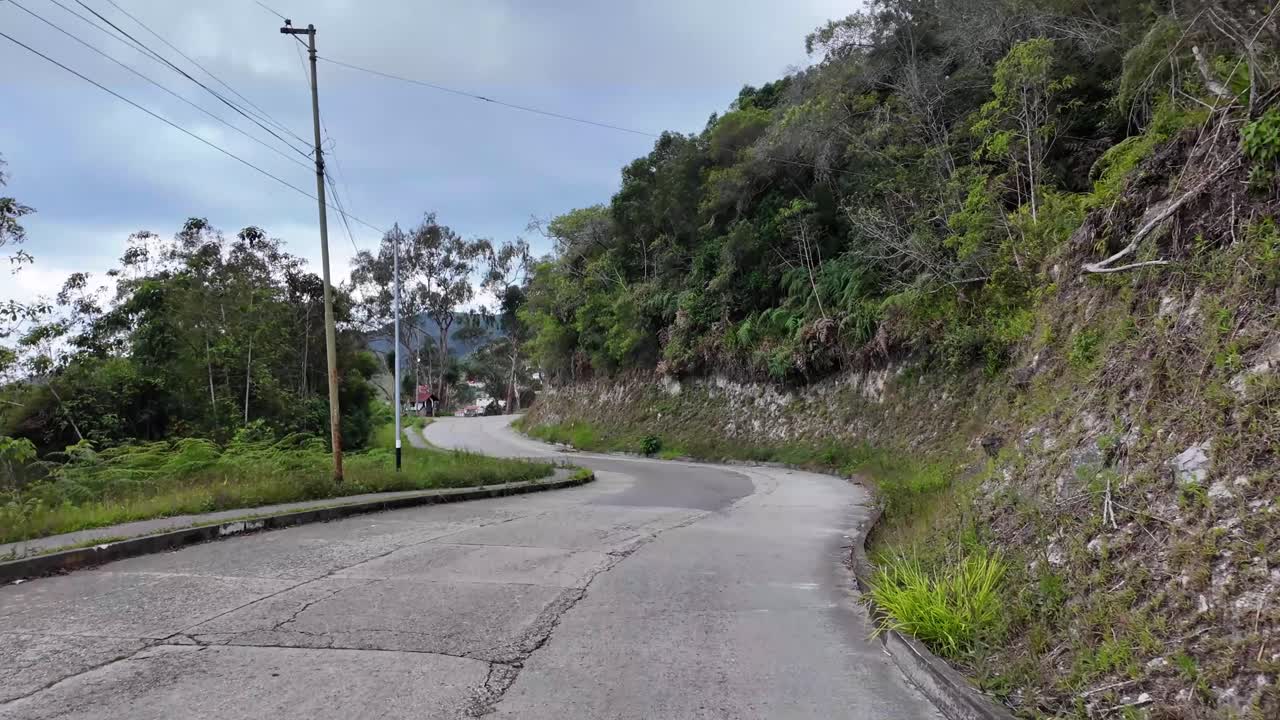POV moviéndose por una exuberante carretera de montaña de hormigón que se abre a la vista de la entrada de una tranquila comunidad de las tierras altas, Venezuela