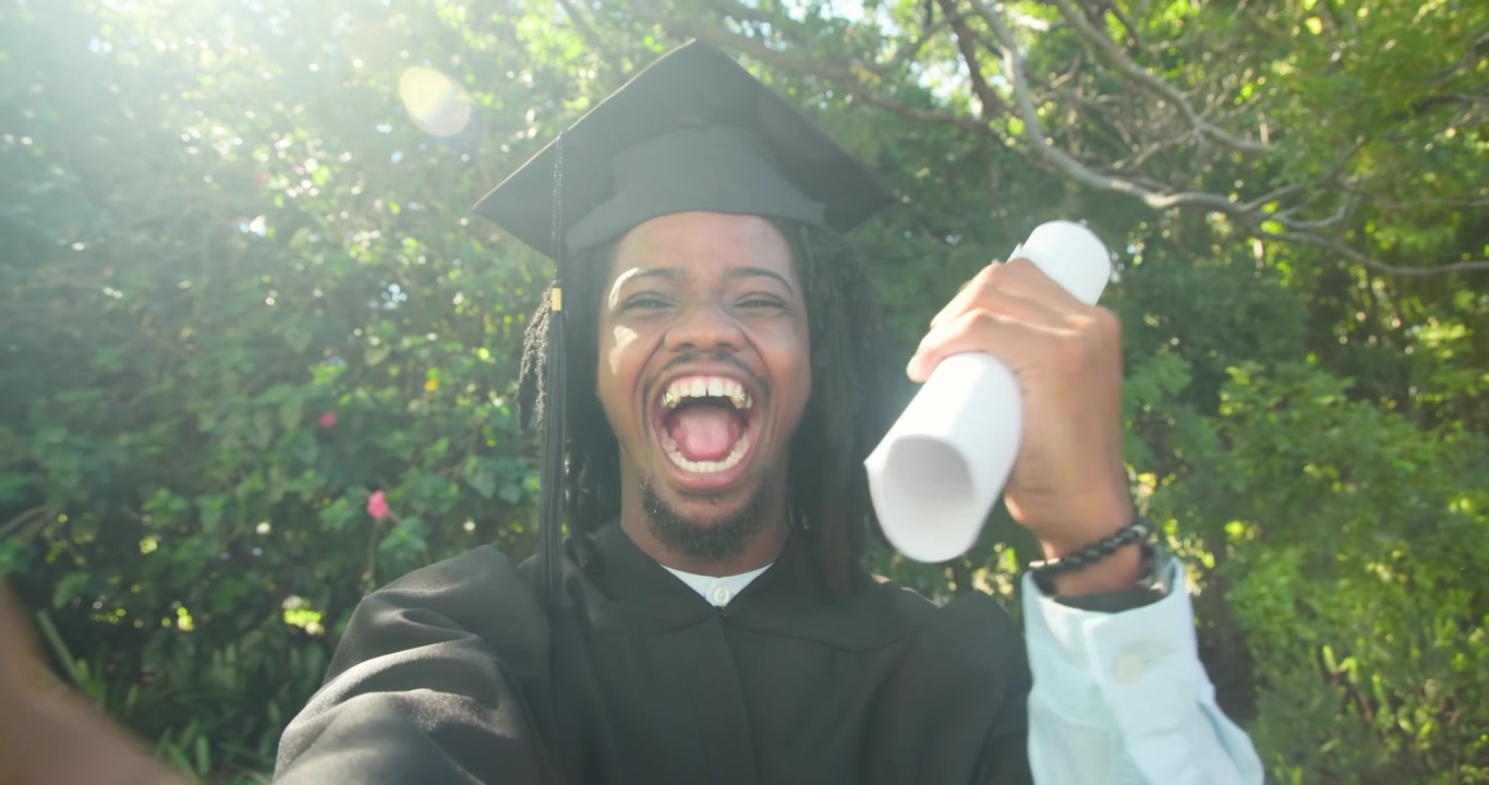 African American man in gown holding diploma celebrating graduation shaking then speaking in garden
