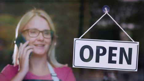 woman talking on the phone standing behind a glass door with a sign open