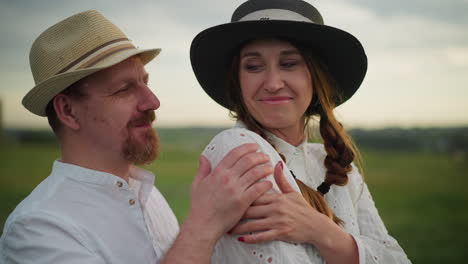 a woman in a hat and white dress smiles contentedly as a man, wearing a brown hat and white shirt, lovingly embraces her from behind in a picturesque countryside setting