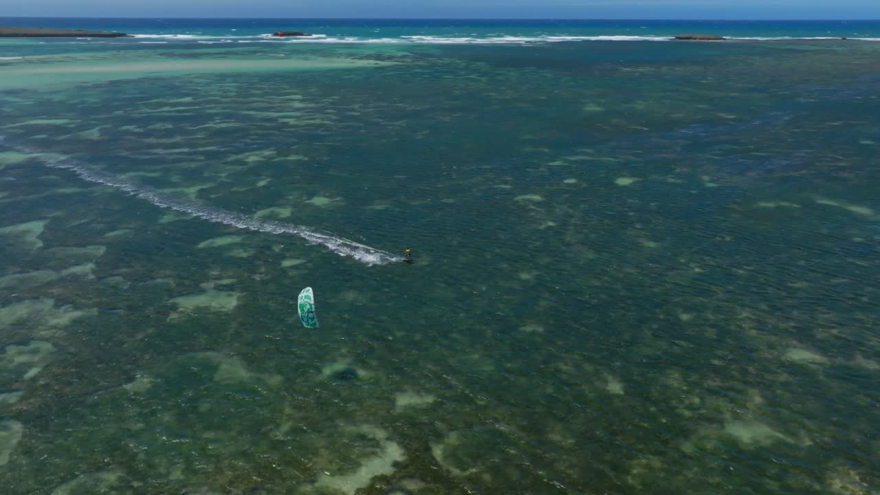 Aerial view as drone follows a kitesurfer above reef waters. It overtakes the rider while tracking movement over the turquoise sea