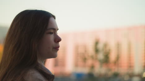 una toma de perfil en primer plano de una joven con una chaqueta de melocotón, caminando por un parque. la toma captura su expresión enfocada y cabello largo, con un fondo urbano borroso