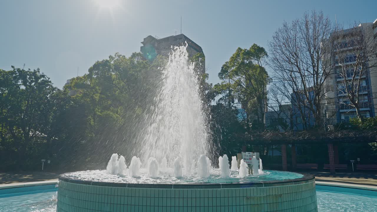 Una toma de primer plano dinámica de agua pulverizándose a lo alto desde la fuente central en el Parque Gyosen, con destellos de sol y árboles verdes al fondo