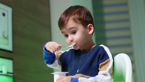 beautiful caucasian boy sits at the chair and eats yogurt. lovely kid enjoys dairy at home. low angle view.