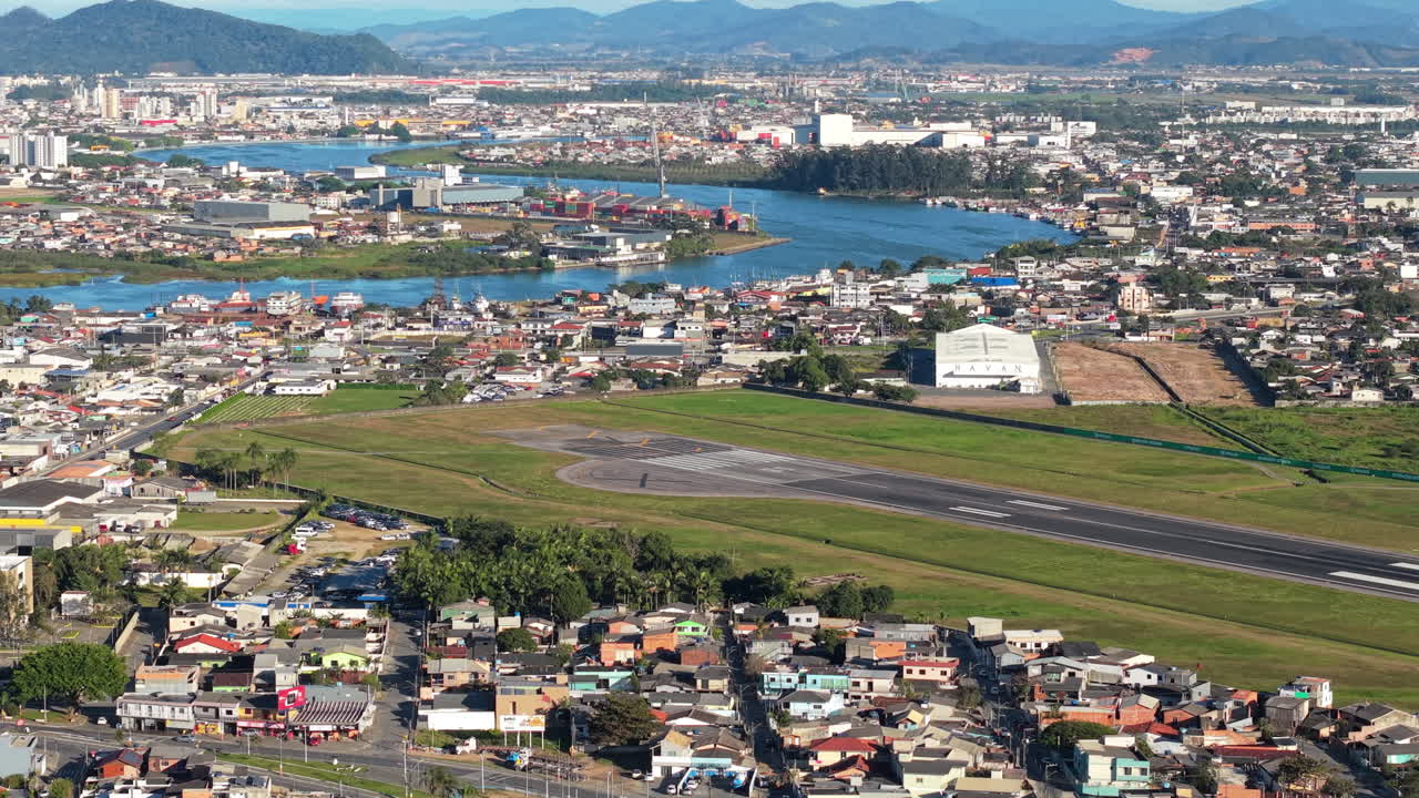 Aerial panorama of Navegantes International Airport, Santa Catarina, Brazil, showing runway, urban neighborhoods, and the Itajaí River with the city of Itajaí in the background under a clear sunny sky