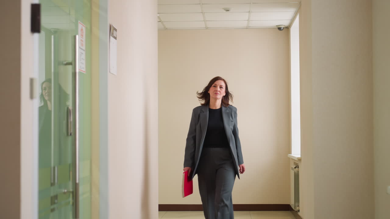 Confident businesswoman in dark formal suit walking down modern office hallway holding red folder, focused and professional, suggesting arrival at meeting or important appointment in corporate setting