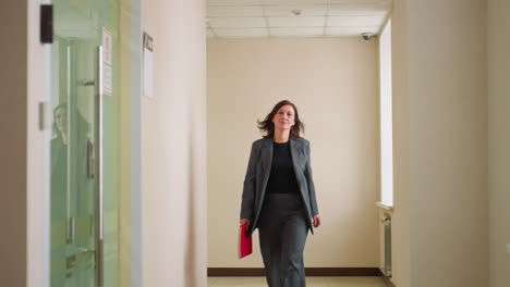confident businesswoman in dark formal suit walking down modern office hallway holding red folder, focused and professional, suggesting arrival at meeting or important appointment in corporate setting